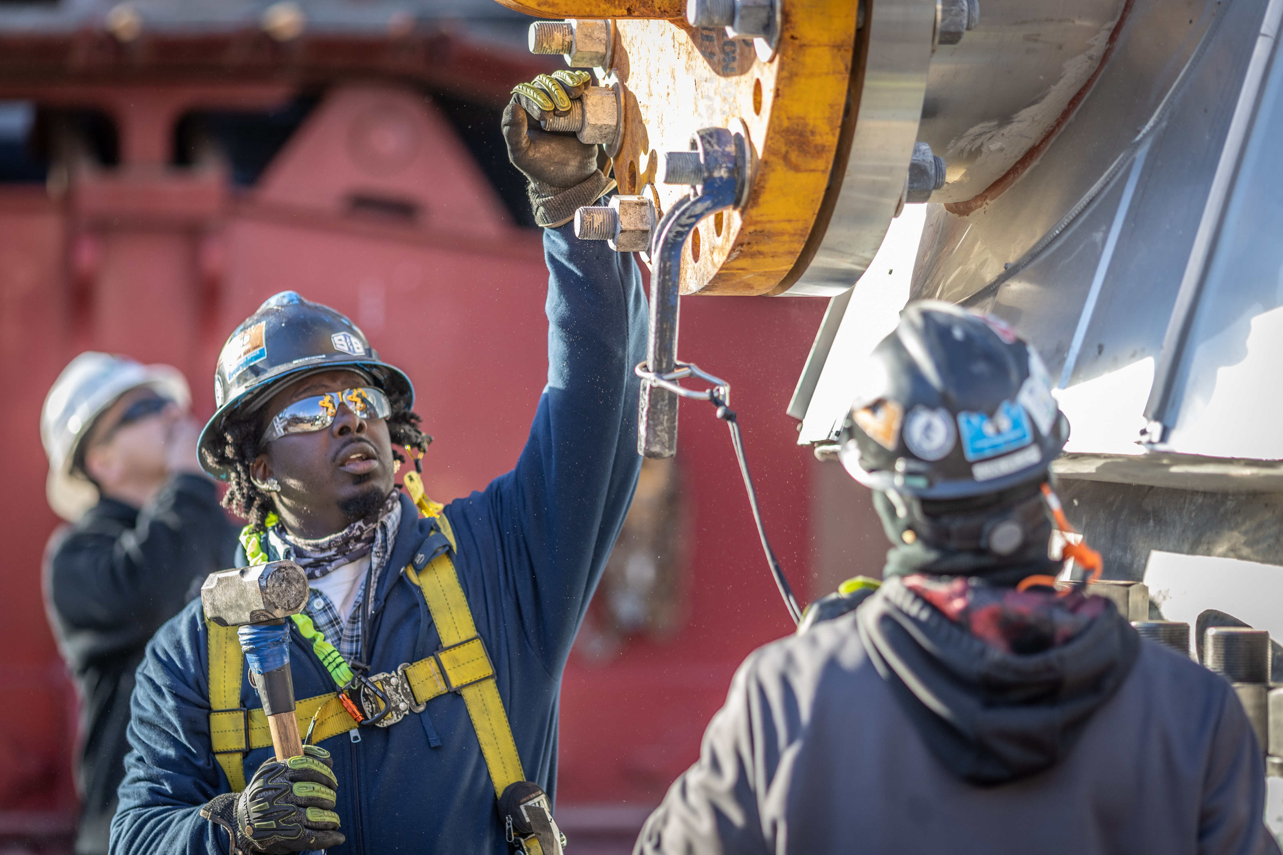 Man with sunglasses and a hard hat reaching up to a large industrial gear