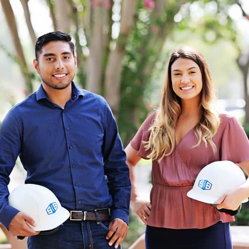 Two young professionals smiling at the camera holding hardhats